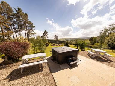 An outdoor area with a covered hot tub two white picnic tables gravel and stone flooring with trees and hills in the background at Heatherbrae in Middleton-In-Teesdale