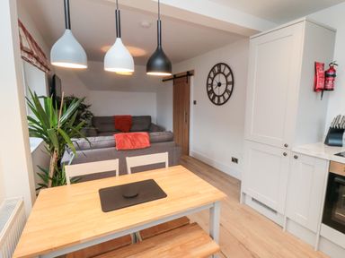 A dining area with a wooden table and chairs next to a living room with gray sofas and a large wall clock at 4 Maypole Green in Fylingthorpe