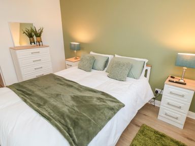 A bedroom with a bed with green pillows and throw a white chest of drawers with plants and two bedside tables with lamps at 4 Maypole Green in Fylingthorpe