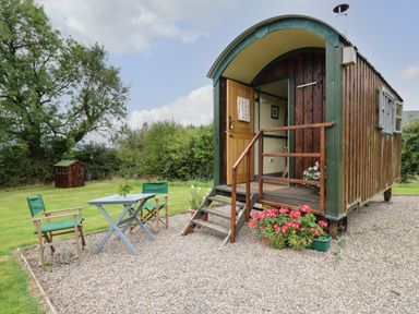 A shepherds hut with steps and flowers outside and a table with two chairs on gravel at Brown Hare Shepherds Hut in Llangorse