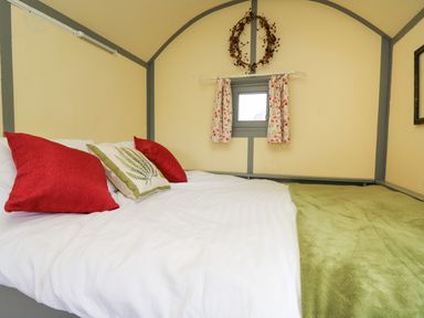 A bedroom with a bed with red and green pillows a small window with floral curtains and a wreath on the wall at Brown Hare Shepherds Hut in Llangorse