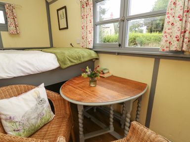 A small room with a wooden table holding a vase of flowers and books next to a bed with a green blanket and wicker chair with a rabbit pillow by a window at Brown Hare Shepherds Hut in Llangorse