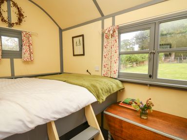 A bedroom with a bed, small windows with floral curtains, a wooden chest with books and a vase of flowers at Brown Hare Shepherds Hut in Llangorse