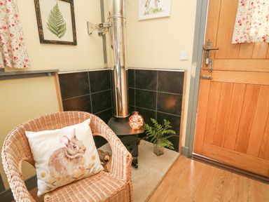 A corner of a room with a wicker chair with a rabbit cushion a wood stove with a metal chimney a wooden door and wall art at Brown Hare Shepherds Hut in Llangorse
