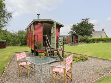 A red gypsy-style caravan with open door on a gravel area with table and two striped chairs at Tilly Gypsy-style Caravan Hut in Llangorse