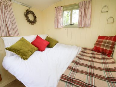 A bed with white sheets green and red pillows a plaid blanket and a window with striped curtains in a small room at Tilly Gypsy-style Caravan Hut in Llangorse