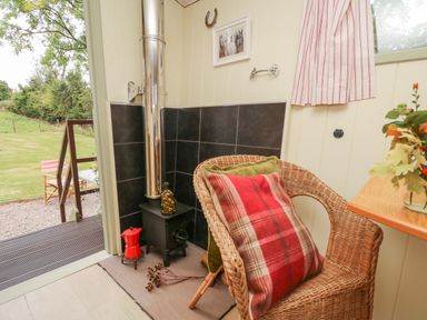A corner with a wicker chair holding pillows next to a small wood stove with a metal chimney pipe at Tilly Gypsy-style Caravan Hut in Llangorse