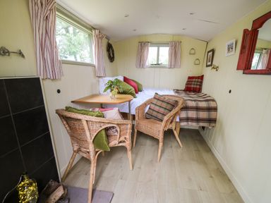 A small room with two wicker chairs a wooden table and a bed with cushions and blankets at Tilly Gypsy-style Caravan Hut in Llangorse