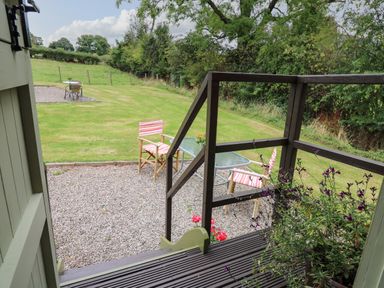 A garden view with striped chairs and a glass table on gravel near a wooden deck and grass at Tilly Gypsy-style Caravan Hut in Llangorse