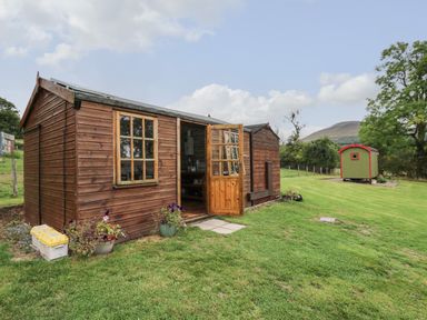 A wooden shed with an open door on a grassy lawn with a smaller green hut in the background at Tilly Gypsy-style Caravan Hut in Llangorse