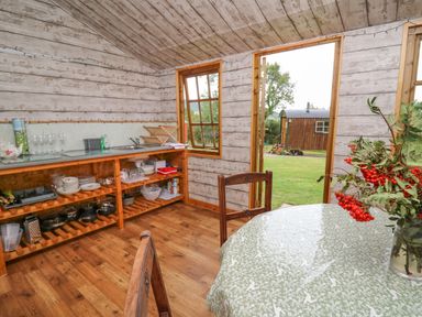 A rustic kitchen area with open shelves containing dishes and pots a table with a floral tablecloth and red berries in a vase and an open door showing a grassy yard and another wooden structure at Tilly Gypsy-style Caravan Hut in Llangorse