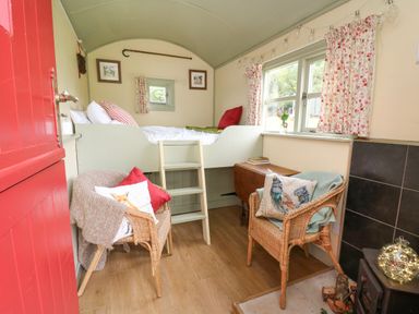 A small room with a red door two wicker chairs a bed with a ladder wooden floor and floral curtains at Sweet Briar Shepherds Hut in Llangorse