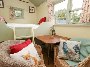 A small room with a raised bed with a small ladder a wooden table with a vase of flowers and two wicker chairs with animal print cushions at Sweet Briar Shepherds Hut in Llangorse