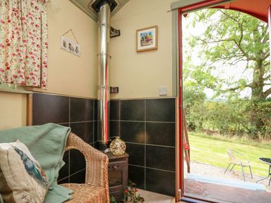 A small seating area with a wicker chair and wood stove next to an open door showing a garden at Sweet Briar Shepherds Hut in Llangorse