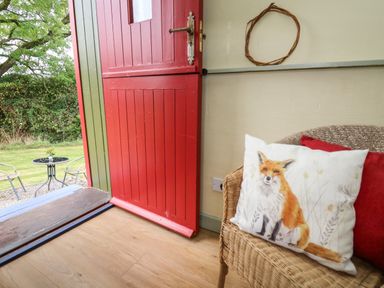 A wicker chair with fox and red cushions next to a partially open red and green door showing an outdoor table and chairs at Sweet Briar Shepherds Hut in Llangorse