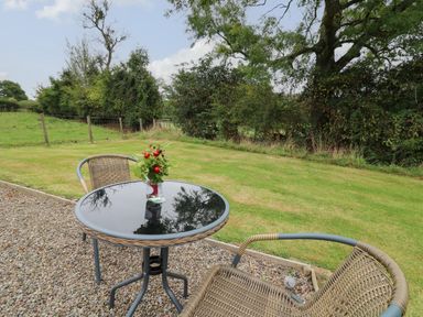 A small outdoor table with a vase of flowers and two wicker chairs on a gravel patio overlooking a grassy yard with trees at Sweet Briar Shepherds Hut in Llangorse
