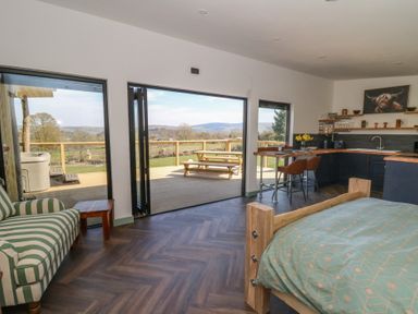 A room with a bed a striped armchair a kitchen area a wooden dining table and an open glass door leading to an outdoor deck with a picnic table at Bryn Hafod in Llandrindod Wells