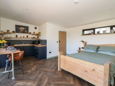 A studio apartment with a wooden bed and green bedding a kitchen with dark cabinets and wooden countertops and a wooden dining table with chairs at Bryn Hafod in Llandrindod Wells