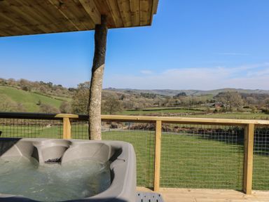 A hot tub on a wooden deck with a railing overlooking green fields and hills at Bryn Hafod in Llandrindod Wells