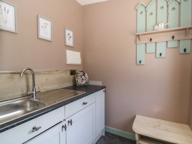 A utility room with a sink white cabinets a wooden bench and wall hooks with framed pictures on the wall at Llety Cariad in Llandrindod Wells