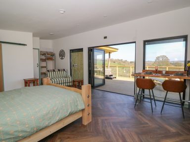 A bedroom with a wooden bed and a green patterned duvet a striped armchair small wooden tables and a high dining table with two chairs near windows and an open door leading to a wooden deck with countryside views at Llety Cariad in Llandrindod Wells