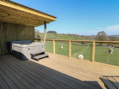 An outdoor wooden deck with a hot tub and a railing overlooking a green field at Llety Cariad in Llandrindod Wells
