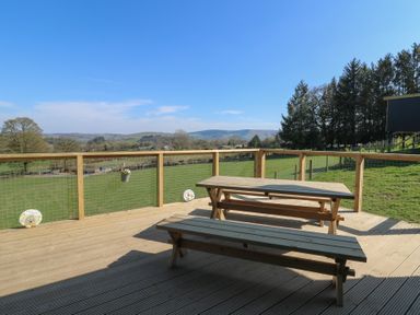 A wooden outdoor deck with picnic tables and a view of green fields and trees at Llety Cariad in Llandrindod Wells