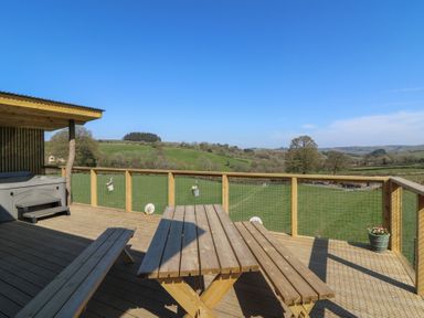 A wooden deck with a picnic table and benches and a hot tub overlooking green fields and hills at Llety Cariad in Llandrindod Wells