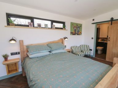 A bedroom with a wooden bed, patterned bedding, two striped chairs, small plants, and a door leading to a bathroom at Llety Cariad in Llandrindod Wells