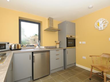 A kitchen with gray cabinets a small refrigerator a microwave a toaster a built-in oven a wooden chair and a wall clock at Lillemor in South Tawton near Whiddon Down