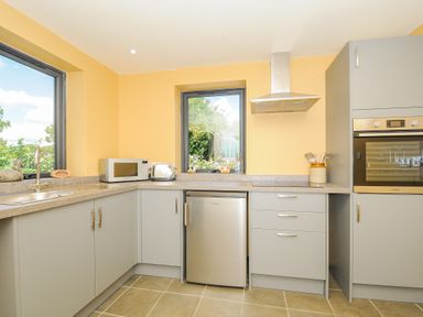 A kitchen with gray cabinets yellow walls a microwave a toaster a small refrigerator a built in oven a stainless steel sink and two windows at Lillemor South Tawton near Whiddon Down
