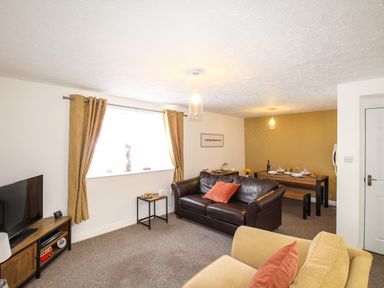A living room with a brown leather sofa, beige armchairs, a wooden TV stand, and a dining table with tableware at Brewers Retreat in Weymouth