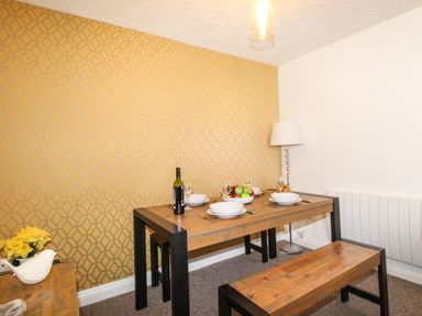 A dining area with wooden table and benches set with white plates wine glasses a bottle of wine fruit bowl and a floor lamp against patterned yellow and white walls at Brewers Retreat in Weymouth