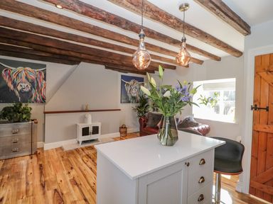 A kitchen island with a vase of flowers and two hanging pendant lights in a room with wooden ceiling beams a brown wooden door and a red sofa with cushions at O'Neil Cottage Beadnell