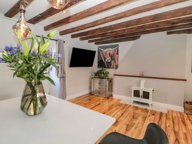 A living room with wooden beams on the ceiling a wall-mounted television a colorful painting a small white stove and a glass vase with flowers on a counter at O'Neil Cottage in Beadnell