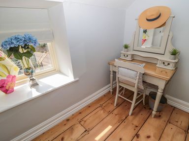 A small vanity desk with a mirror a chair with a cushion and a hat hanging above it beside a window with flowers on the sill at O'Neil Cottage in Beadnell