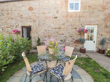 A stone wall exterior with a glass table and four wicker chairs on a stone patio surrounded by plants and flowers at O'Neil Cottage Beadnell