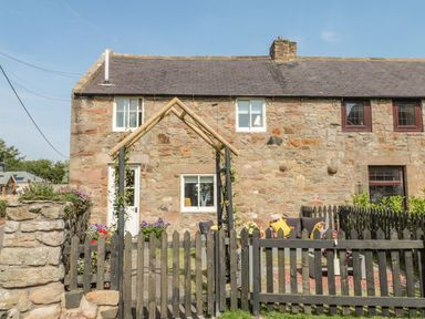 A stone cottage with a black picket fence and seating area with cushions in the garden at O'Neil Cottage in Beadnell