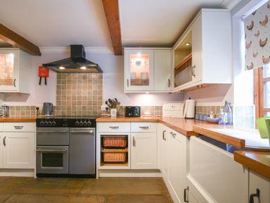 A kitchen with white cabinets wooden countertops a stove a toaster and a window with chicken patterned blind at Barley Cottage Weycroft near Axminster