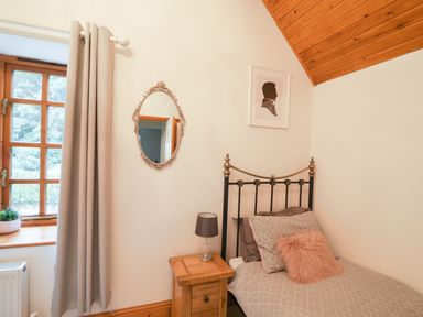 A bedroom with a single bed beside a wooden nightstand with a lamp and a window with grey curtains at Barley Cottage in Weycroft near Axminster
