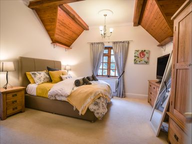 A bedroom with a bed bedside tables a window with curtains a chest of drawers a standing mirror and wooden ceiling panels at Barley Cottage Weycroft near Axminster