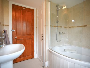 A bathroom with a bathtub and shower, a wooden door, a white sink, and beige tiled walls at Barley Cottage in Weycroft near Axminster