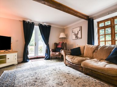 A living room with a beige sofa black cushions an armchair with a lamp near a deer painting a wooden cabinet with a TV blue curtains and a patterned rug at Barley Cottage Weycroft near Axminster