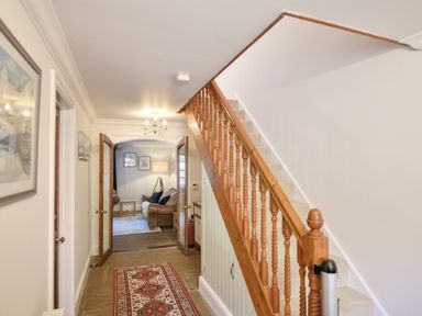 A hallway with wooden staircase and patterned rug leading to a living room with brown sofa at Barley Cottage in Weycroft near Axminster