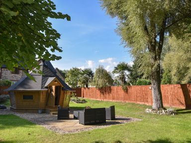 A garden with a wooden cabin a seating area with black chairs and a large tree at Barley Cottage in Weycroft near Axminster