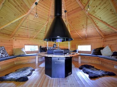 An octagonal wooden room with a central black grill and chimney surrounded by built in benches with cushions and fur rugs on the floor at Barley Cottage in Weycroft near Axminster