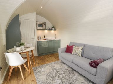 A small living space with a grey sofa, a round dining table with two chairs, and a kitchenette with green cabinets at Yr Onnen in Bontuchel near Ruthin