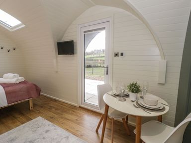 A small room with a bed with towels on it a round dining table set for two with dishes glasses and a plant a wall mounted TV and a door showing an outdoor view at Yr Onnen in Bontuchel near Ruthin