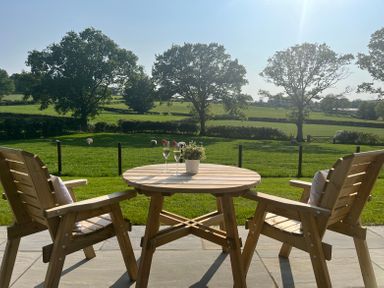 A wooden table with two glasses of drink and a potted plant between two wooden chairs on a patio overlooking a green field with trees and sheep at Yr Onnen in Bontuchel near Ruthin