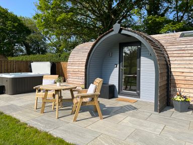 A wooden pod with a glass door on a stone patio with a round wooden table two wooden chairs and a hot tub in the background at Yr Onnen in Bontuchel near Ruthin